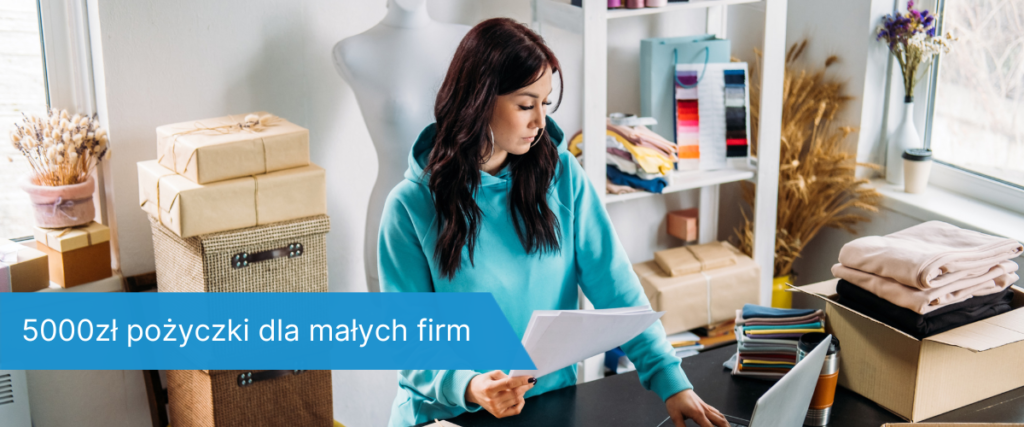 Młoda kobieta w niebieskiej bluzie stoi przy biurku z dokumentami w ręku, otoczona paczkami i komputerem